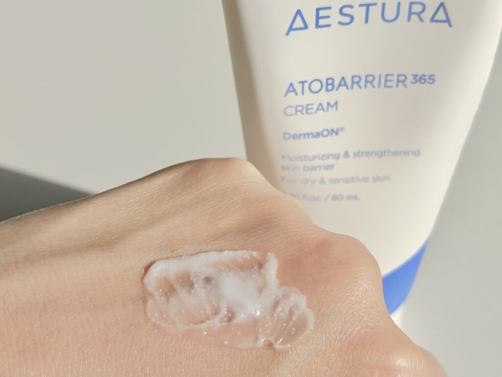 Aestura Atobarrier 365 Cream swatched on the back of a hand revealing visible white DermaON ceramide capsules next to the product tube