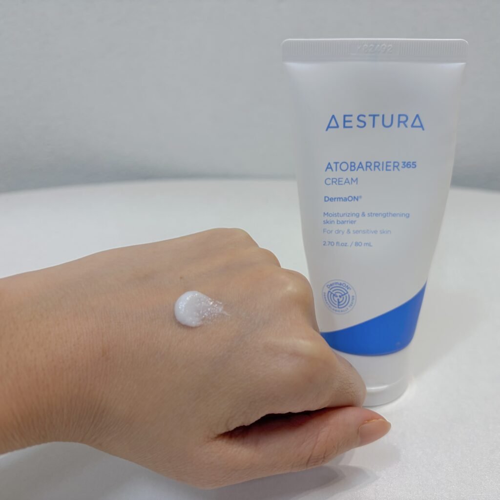 A dollop of Aestura Atobarrier 365 Cream on the back of a hand showing visible DermaON capsules alongside the product tube in the background