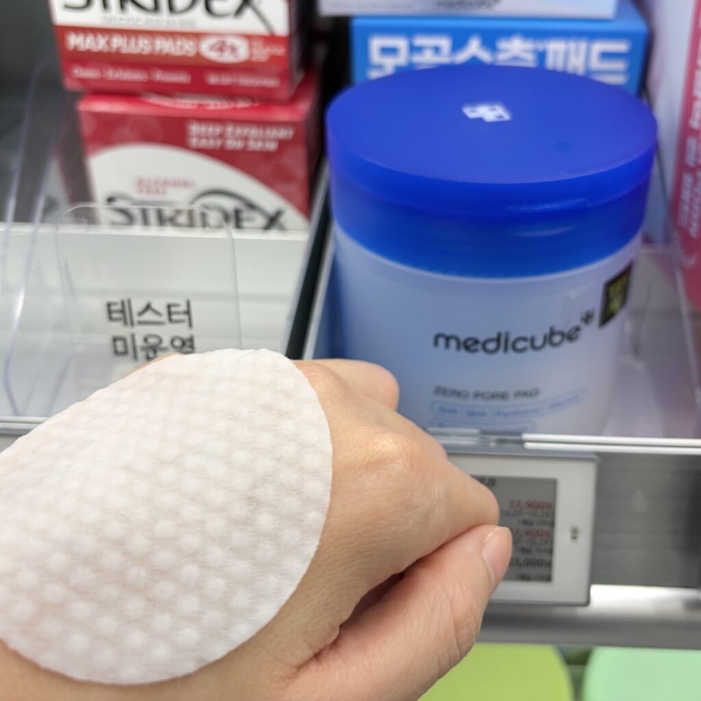 Medicube Zero Pore Pad 2.0 embossed pad placed on the back of a hand with the blue product container displayed in the background at Olive Young